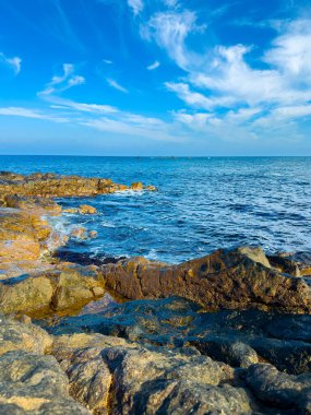Dalgalar bulutlu mavi bir gökyüzünün altında kayalık sahile vurur. Güneş suyun üzerinde parlıyor, sakin okyanus dalgalarını ortaya çıkarıyor. Bu sahne gündüz doğayı gösteriyor..