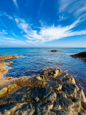 Kayalar okyanusun kenarında bulutlu açık mavi bir gökyüzünün altında. Dalgalar, güneş suyun üzerinde parlarken yavaşça kayalara çarpıyor..