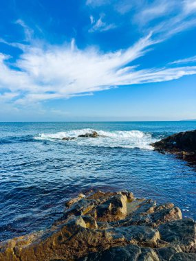 Açık mavi gökyüzü okyanusu kayalara çarpan dalgalarla kaplıyor. Güneş tepede ve su parlak ışığı yansıtıyor. Kıyıda büyük taşlar ve yumuşak dalgalar var..