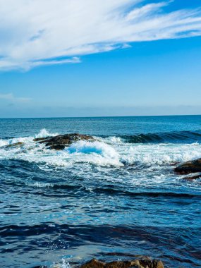 Parlak bulutlar mavi okyanusun üzerinde süzülür. Dalgaların gün ortasında kıyıda kayalara çarptığı yerde. Sahne berrak bir gökyüzünü ve sakin suları gösteriyor..
