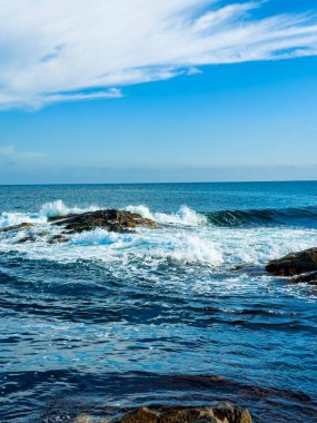 Okyanus dalgaları kıyıdaki kayalara vururken güneş parlıyor. Mavi gökyüzünün birkaç bulutu vardır ve deniz gün ışığında derin ve engin görünür..