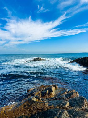 Dalgalar, öğleden sonra kıyı şeridine ulaştıklarında kayaların üzerinden hareket eder. Gökyüzü, ufukta okyanus suyunun üzerinde birkaç bulut olan parlak bir maviyi gösteriyor..