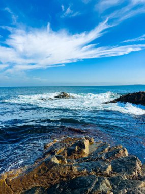 Dalgalar kıyı şeridindeki engebeli kayalara çarparken bulutlar öğleden sonra suyun üzerinde mavi gökyüzünde sürükleniyor. Sahne, okyanusun enerjisini yakalar..