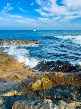 Dalgalar kıyı boyunca kayalara çarparken bulutlar gökyüzünde yüzüyor. Güneş okyanusu aydınlatıyor, kıyı şeridinde parlak bir manzara yaratıyor..