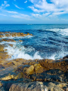 Dalgalar kayalık kıyıya su sıçramalarıyla vurur. Güneş gökyüzünde ışıl ışıl parlıyor. Mavi su ufka kadar uzanıyor. Bulutlar gökyüzünde süzülür.