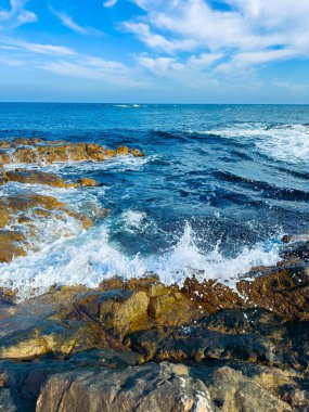 Su kıyı şeridi boyunca kayaların üzerinden akar. Yumuşak dalgalar bulutlu mavi bir gökyüzünün altında kıyıya doğru hareket ediyor. Güneş sahneyi aydınlatıyor..