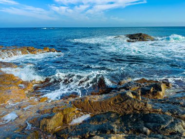 Dalgalar geliyor ve kıyıya yakın kayalara çarpıyor. Güneş yukarıda parlıyor, ve okyanus ufka doğru uzanıyor, doğanın hareketinin bir sahnesini oluşturuyor..