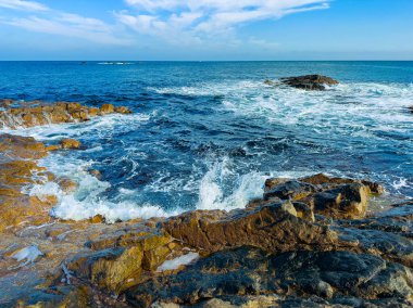 Dalgalar kıyı şeridi boyunca kayalara çarpıyor. Su sıçrıyor ve sular yükseldikçe köpük çıkıyor. Gökyüzü mavi boşluklu bulutları gösteriyor. Bu sahne gün ışığında geçiyor..