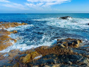 Okyanus dalgaları kıyı boyunca kayalara çarpıyor. Deniz parlak mavi bir gökyüzünün altında uzanır bazı bulutlarla. Bu sahne bir kıyı şeridinin yakınındaki doğanın güzelliğini gösteriyor..