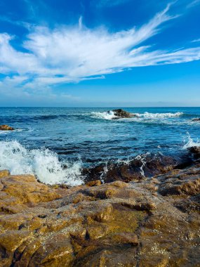 Güneş tepede parlarken dalgalar kıyı boyunca kayalık oluşumlara çarpar. Bulutlar açık mavi gökyüzüne yayıldı ve su kenarında parlak bir manzara yarattı..