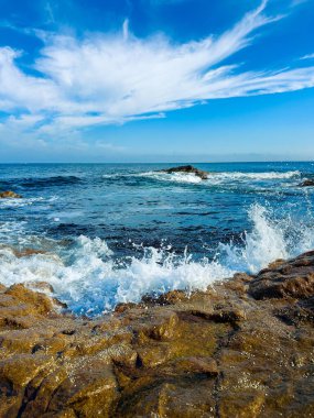 Dalgalar kıyı boyunca parlak bir gökyüzünün altındaki kayalara çarpar. Güneş suyun üzerinde parlıyor ve bulutlar yukarı doğru sürükleniyor. Sahne su ve taşların hareketlerini gösteriyor..
