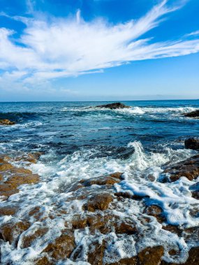 Güneş ışıl ışıl parlarken dalgalar kıyı şeridinde kayaların üzerinde yuvarlanıyor. Deniz, gün ortasında gökyüzünde birkaç bulutla çok uzaklara doğru uzanıyor..