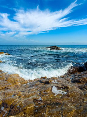 Mavi gökyüzünde bulutlar sürüklenirken dalgalar kıyı şeridi boyunca kayalara çarpıyor. Deniz, parlak güneş ışığı altında uzak mesafelere kadar uzanıyor. Bu sahne doğanın elementlerinin birlikte çalıştığını gösteriyor..