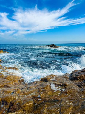Dalgalar kıyı boyunca kayalara çarpıyor bulutlu mavi bir gökyüzünün altında. Su berrak ve güneş ışığını yansıtıyor. Sahne, su ve kayalarla dolu bir kıyı bölgesini gösteriyor..