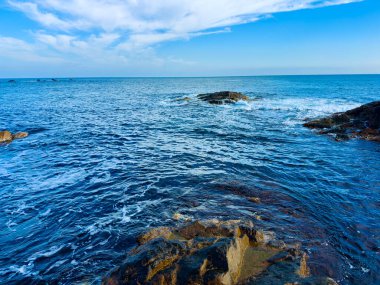 Dalgalar kıyı şeridi boyunca kayalara doğru ilerliyor. Uçsuz bucaksız okyanus ufka uzanır parlak mavi gökyüzünün altında bulutlarla.