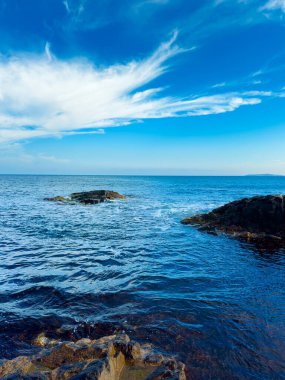 Parlak mavi okyanus ufka doğru uzanır ve kıyıda kayalar görülebilir. Gökyüzü bulutlardan ve gün boyunca güneş ışığını gösteren berrak bir manzaradan oluşur..