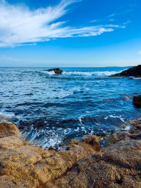 Güneş tepemizde parlarken dalgalar okyanus yakınlarındaki kayaların üzerinden akar. Su ufka kadar uzanıyor, mavi gökyüzü ile birkaç bulutun buluştuğu yere..