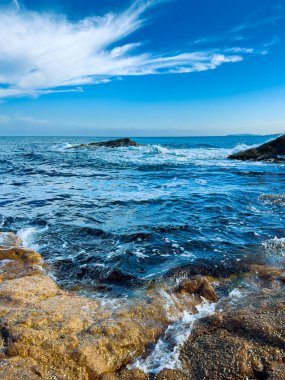 Kıyı bölgesinde dalgalar dalgalanırken su kayalara sıçrıyor. Sahne engin bir okyanusu parlak bir gökyüzünün altında bazı bulutlarla gösteriyor..
