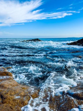 Dalgalar güneş ışığına çarptığında su kıyı boyunca kayaların üzerinden akar. Gökyüzü bulutlarla mavidir, okyanus kenarında canlı bir manzara yaratır..