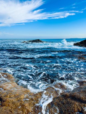 Kıyı şeridinde dalgalar akarken su kayalara çarpıyor. Gökyüzü mavi ve öğleden sonra ışığı altında bulutlar var..