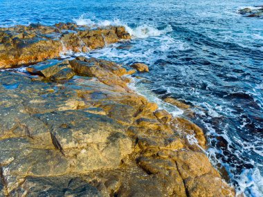 Dalgalar yüzeye vurduğunda su kıyı şeridi boyunca kayaların üzerinden akar. Sahne gündüzleri mavi gökyüzüyle çekiliyor..
