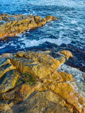 Kıyı şeridinde kayalar görülebilir. Üzerlerinden su akar. Dalgalar taşlara çarpar ve köpük oluşturur. Güneş sahnede parlıyor..