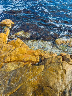 Dalgalar açık gökyüzünün altındaki kayalık bir kıyı şeridine çarpar. Su, sahil boyunca uzanan sert kayalarla buluştuğunda güneş ışığını yansıtır. Bu sahne doğanın elementlerinin güzelliğini yakalar..
