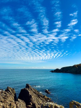 Bulutları sakin okyanus sularının üzerinde uzanan parlak mavi gökyüzü. Rocky sahili denizle kesişiyor ve doğal bir alan yaratıyor. Sahne gündüz saatlerinde geçiyor..