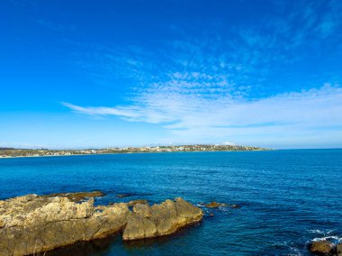 Açık mavi deniz manzarası gündüz boyunca kayalık kıyı şeridi ile buluşuyor. Kıyı boyunca uzak binalar görülür, manzaralı bir manzara yaratır..