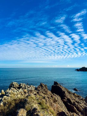 Dalgalar kıyı boyunca kayalara hafifçe çarpar. Gökyüzü bulutları sergiler. Sahne karanın ve denizin planlarını parlak gündüz ışığı altında yakalıyor..