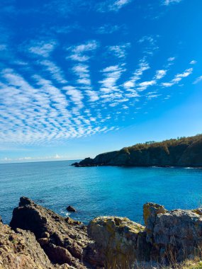 Dalgalar kıyı boyunca kayalara yavaşça vururken bulutlar mavi bir okyanusun üzerinde gökyüzünde ilginç desenler oluşturuyor. Toprak güneşin altında yükselir..
