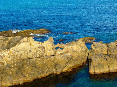 Kayalar açık mavi bir gökyüzünün altındaki kıyı bölgesinde suyun üzerinde görülebilir. Kayaların etrafında hareket eden yumuşak dalgalarla okyanus sakin..