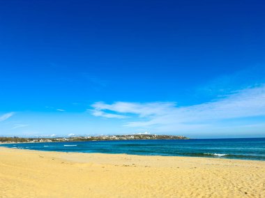 Sahne sahil boyunca uzanan geniş kumlu bir sahili gösteriyor. Sahilde dalgalar yavaşça dolaşırken arka planda açık mavi bir gökyüzünün altında bir kıyı kasabası görülüyor..