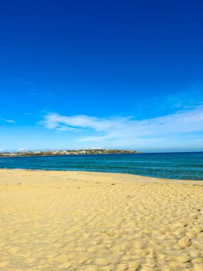 Kumsal açık mavi su boyunca uzanır parlak bir gökyüzü ile. Uzakta, kara güneş ışığı altında görülebilir. Sahne gündüzleri doğal bir ortam gösteriyor..