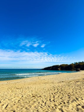 Kumsal, kıyıya vuran yumuşak dalgalarla dolu. Gökyüzü açık mavi ve berrak. Kıyı boyunca güneş ışığı altında kayalar görülebilir..