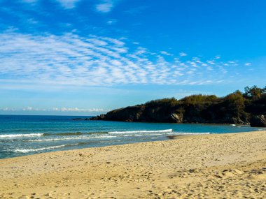 Kumsalda küçük dalgalar kıyıya vururken parlak güneş ışığı parlıyor. Mavi gökyüzü bulutları uzaktaki kayalık bir sahil şeridine dağıttı..