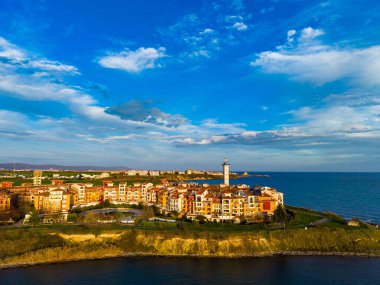 Bir kıyı kasabası kıyıya yakın çeşitli binalara ve bir deniz fenerine sahiptir. Sahne, öğleden sonra bulutlu denizi ve gökyüzünü gösteriyor..