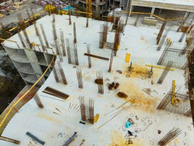 İşçiler inşaat sürecine beton bir levha üzerinde devam ediyorlar. Çelik parmaklıklar sitenin her tarafına yerleştirilmiş. Araçlar ve malzemeler etrafa saçılmış..