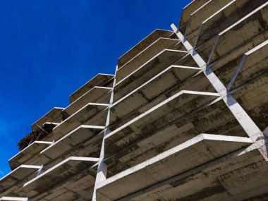 İşçiler birkaç katlı beton bir yapı inşa ediyorlar. Gökyüzü mavi ve berrak. Fotoğraf da destek kirişlerini ve binanın tepesini alçak bir açıdan gösteriyor..