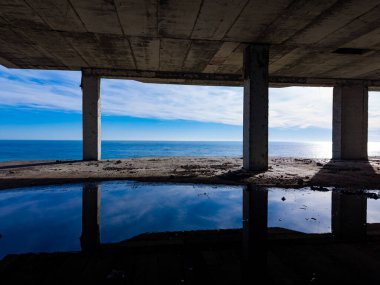 Dalgalar, beton sütunları olan bir yapının altında yavaşça kıyıya çarpar. Bulutlar üstümüzde sürüklenirken içeride su birikiyor. Okyanus gökyüzünün renklerini yansıtır..