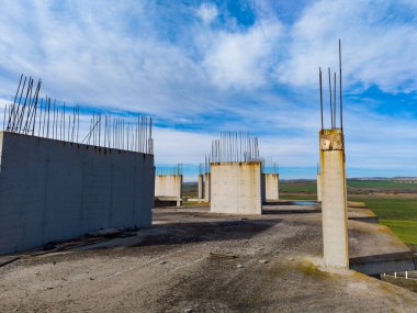 Bitmemiş bir inşaatta beton sütunlar gökyüzüne yükseliyor. Alan, arka planda geniş mavi gökyüzünün altında görünen çimen ve arazi gibi ihmalkârlık belirtilerini gösteriyor..