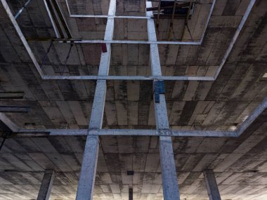 İşçiler beton kirişler ve sütunlarla bir yapı inşa ediyorlar. Metal çubuklar tavanda görünür durumda, bu da devam eden çalışmayı gösteriyor. Güneş ışığı sahneyi aydınlatır..