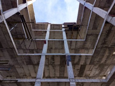 İşçiler, bitmemiş bir binanın uzun ve beton duvarlı olduğu inşaat sahasının etrafında hareket ediyorlar. Açık gökyüzü aşağıdan görülebiliyor, çalışmaların gidişatını gösteriyor.