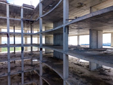 İnşaat alanı açık zemini ve beton duvarları olan bir bina gösteriyor. Boş pencerelerden içeri güneş ışığı giriyor. Toprağı toz ve enkaz kaplıyor..