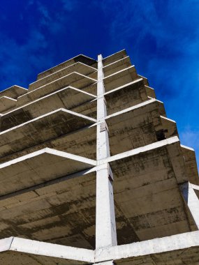 Mavi bir gökyüzünün altında uzun bir beton yapı duruyor. Binanın birkaç katı var. Manzara yerden yukarıya bakıyor. Binanın tasarımı ve yüksekliğini gösteriyor..