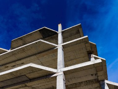 İnşaat halindeki beton bina gökyüzüne doğru yükseliyor. Yapıda birkaç düz seviye ve güçlü sütunlar yer alıyor. Sahne berrak mavi gökyüzü ile.