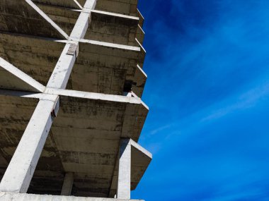 Öğleden sonra mavi gökyüzüne doğru beton bir bina yükselir. Yapının penceresi yok ve iskelesi var. Çevreleyen alanlar açık alan ve inşaat araçları gösteriyor.