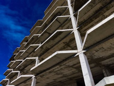 Beton kirişler ve zemin, bir şehirde hala inşa halinde olan bir bina yapısını oluşturuyor. Gökyüzü parlak mavi ve az sayıda bulut sitenin üzerinde görünür..