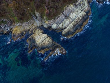 Açık mavi okyanus suyu olan kayalık bir sahil şeridi. Manzara kayalarda seyrek bitki örtüsüne sahiptir. Resim, doğanın güzelliğini havadan yansıtıyor..