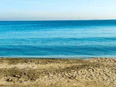 Gün ortasında açık gökyüzü olan bir plajda su kumla buluşur. Dalgalar yavaşça yaklaşırken ufukta görünen uzak gemiler basit bir sahil manzarası yaratıyor..